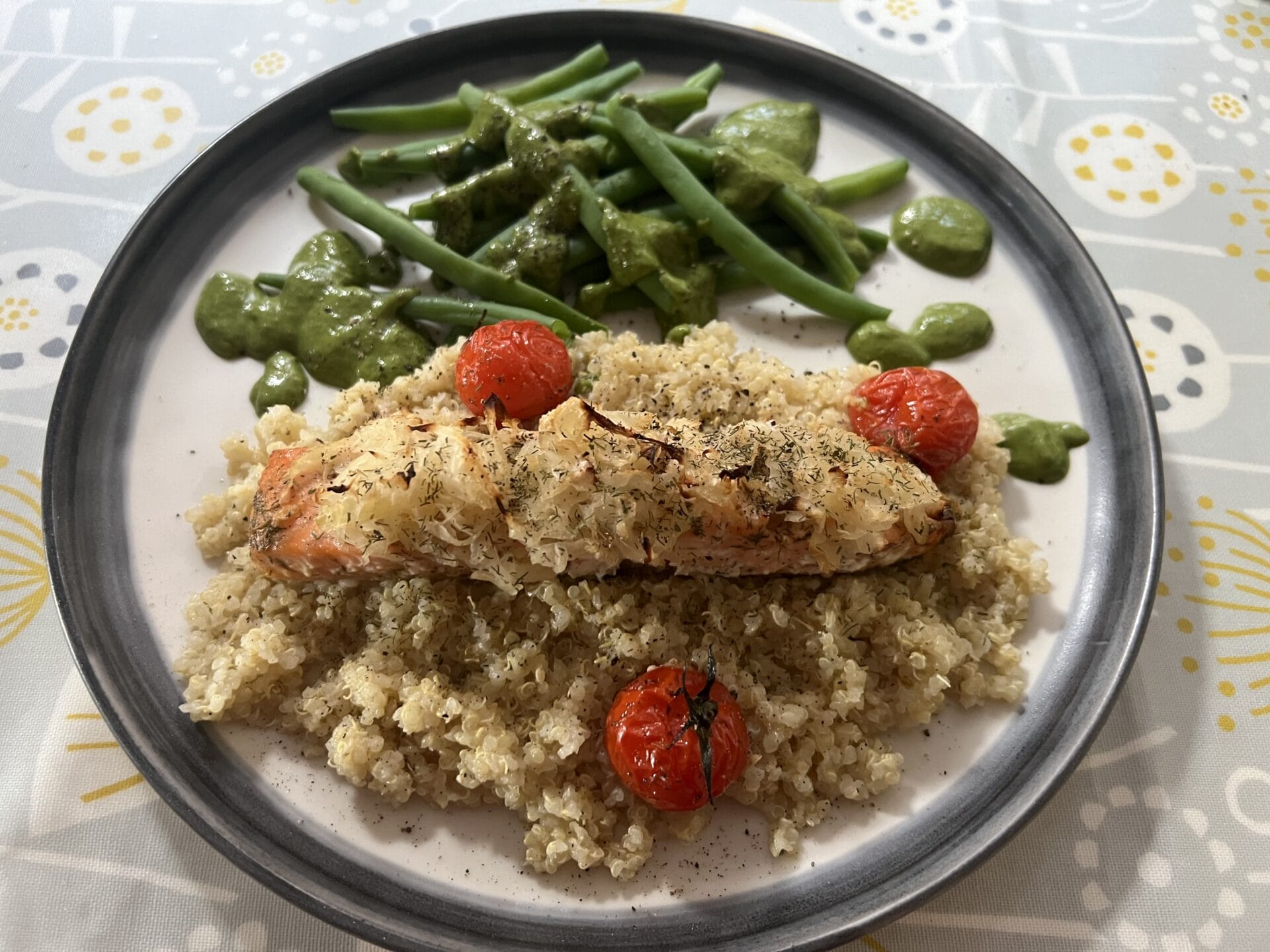 A plate of quinoa topped with oven baked wild salmon and roasted cherry tomatoes, accompanied by green beans with pesto sauce and a side of sauerkraut.