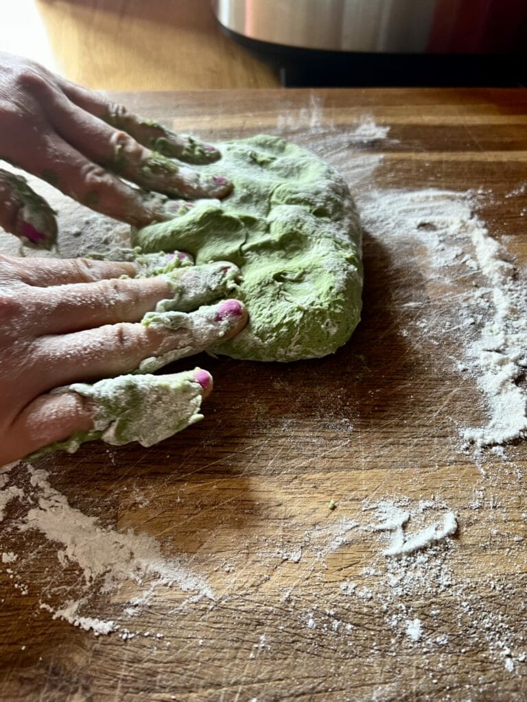 A person's hands are making green pasta dough on a wooden table.