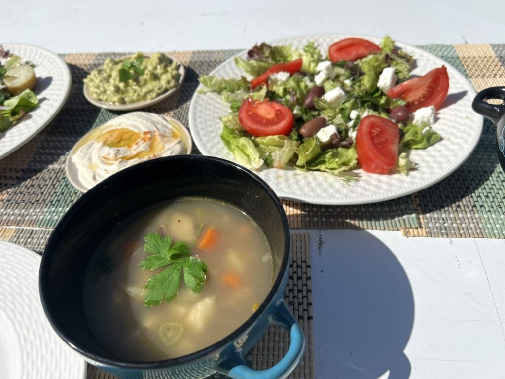 A bowl of soup with a side of Ikarian longevity black-eyed peas cold salad and dips served on an outdoor table.