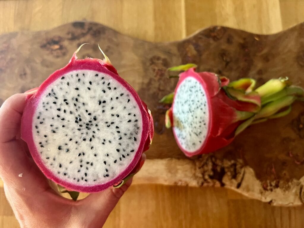 A person holding a sliced dragon fruit, showing the white pulp and black seeds, with another piece on a wooden cutting board.