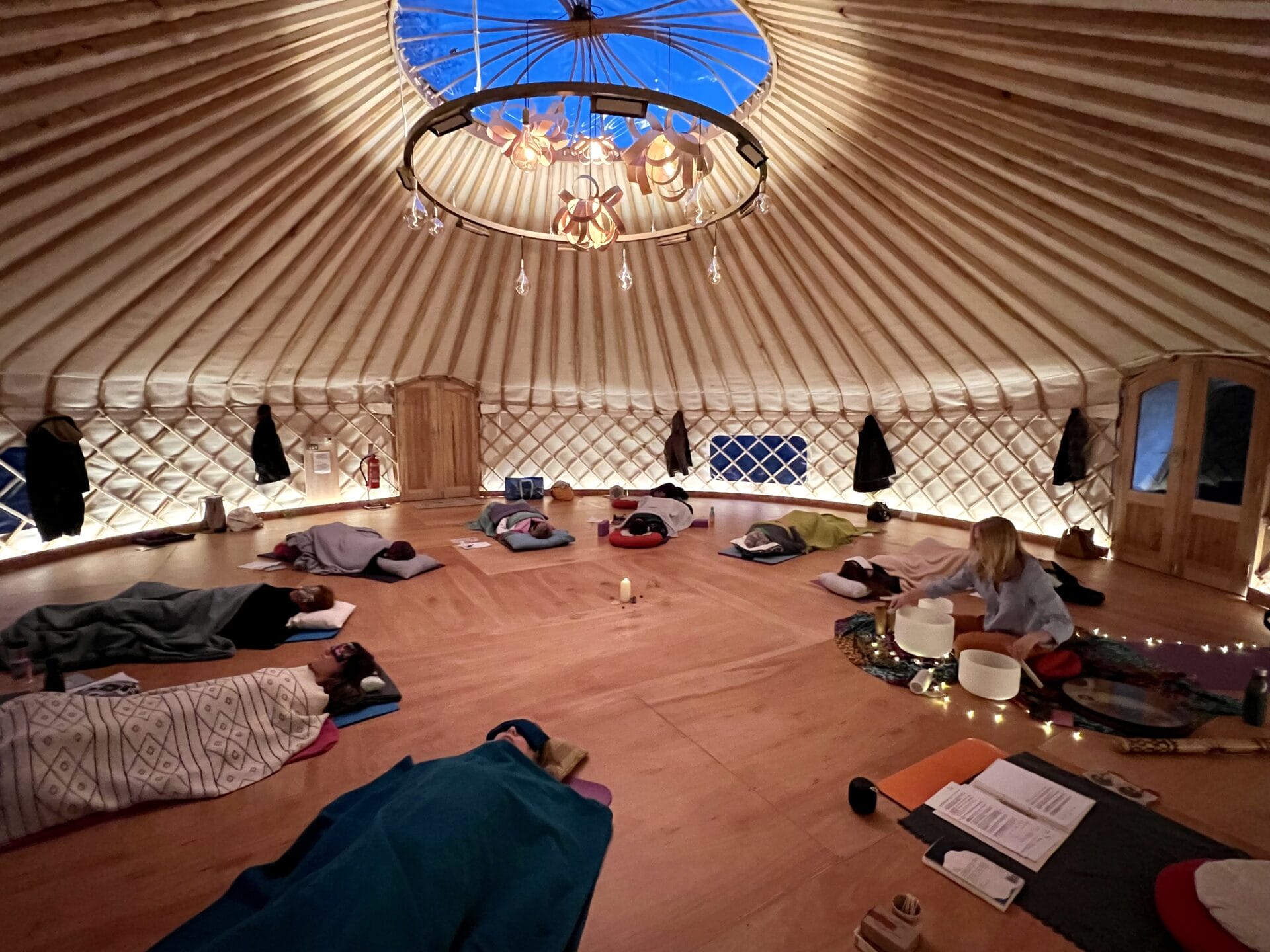 People lie on yoga mats under blankets in a circular wooden yurt with a skylight and soft lighting. A central candle and various items are on the floor. The setting is warm and serene. People lie on yoga mats under blankets in a circular wooden yurt with a skylight and soft lighting. A central candle and various items are on the floor. The setting is warm and serene.