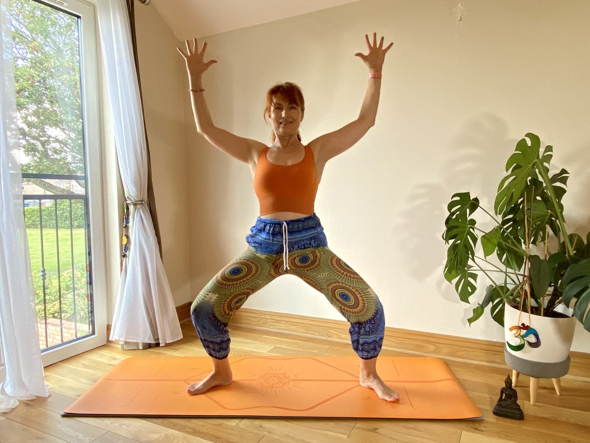 A person stands in a yoga pose on an orange mat in a well-lit room with a large window, wearing a bright orange top and colorful patterned pants. A potted plant is visible in the background.
