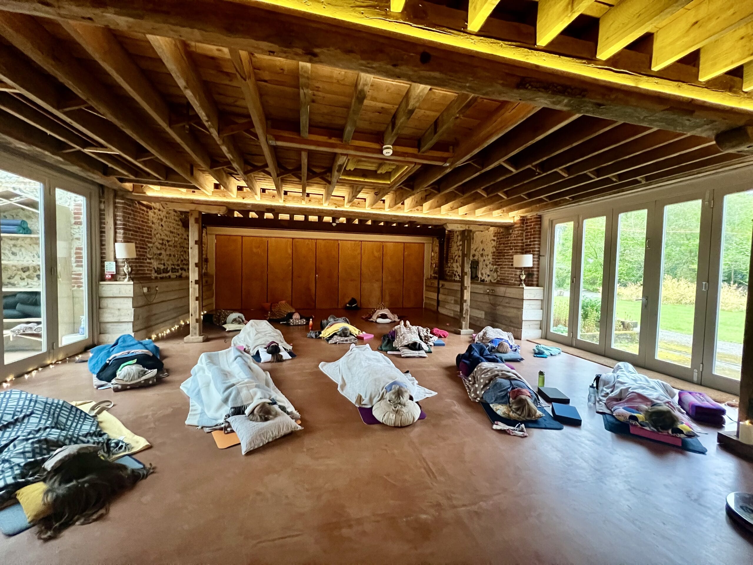 In a spacious room with wooden beams and large windows, a group of people lies on mats under blankets, uniting in mindfulness during our day of shared presence and reflection.