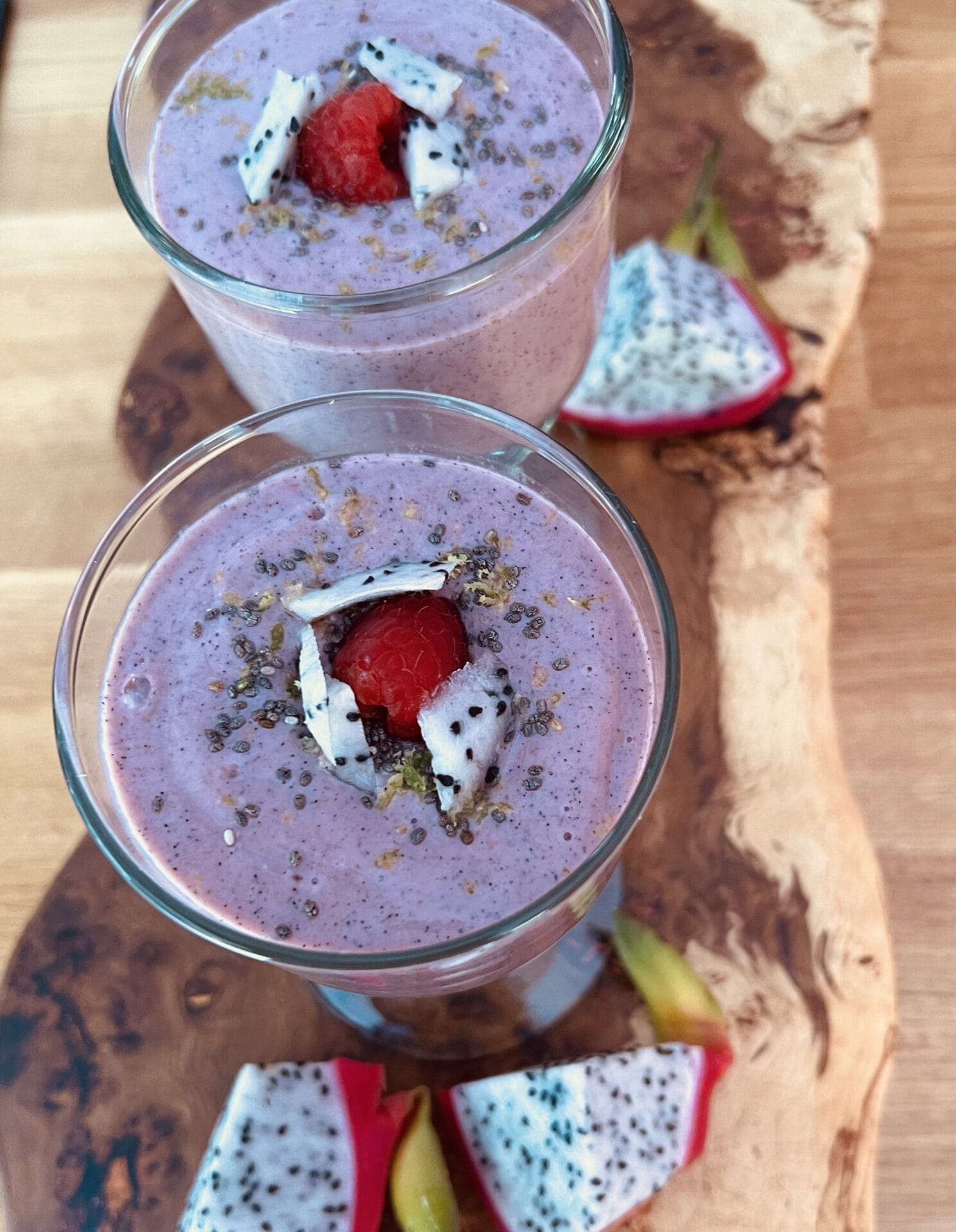 Two glasses of raspberry dragon fruit smoothie topped with chia seeds and fresh fruit, served on a wooden board.