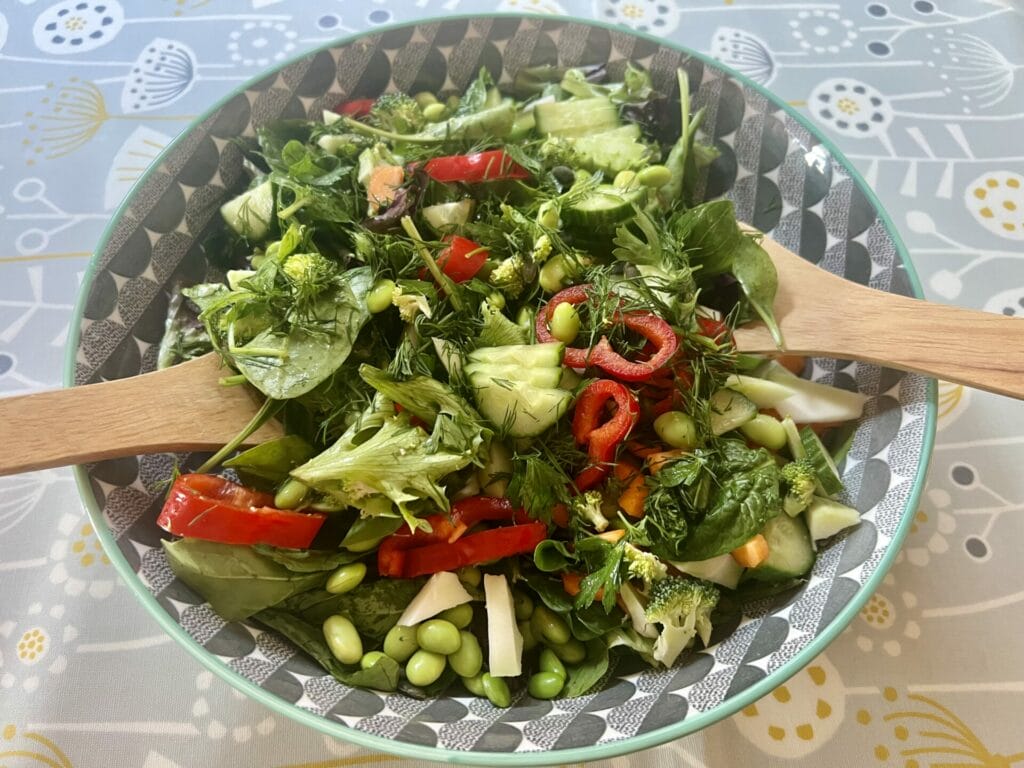 A bowl of fresh Gut Healthy Summer Salad containing a mix of leafy greens, sliced red bell peppers, edamame, quinoa, broccoli, and herbs, with grilled tofu and two wooden salad servers.