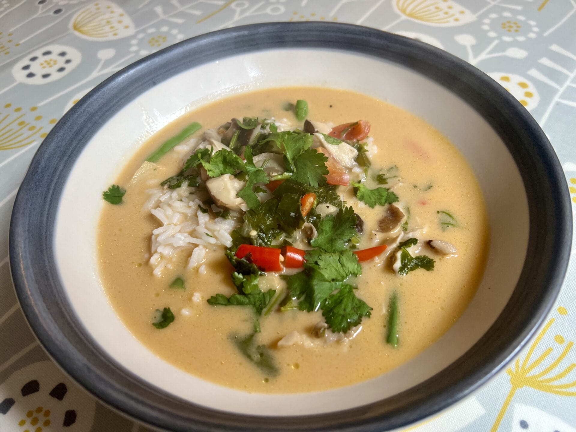A bowl of curry soup with rice, vegetables, mushrooms, and fresh cilantro on top, set on a patterned tablecloth.
