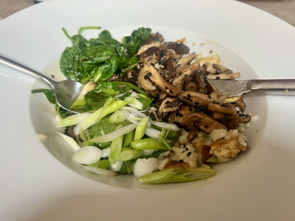 A white bowl filled with sautéed mushrooms, spinach, sliced green onions, and chopped walnuts, accompanied by a spoon and knife.