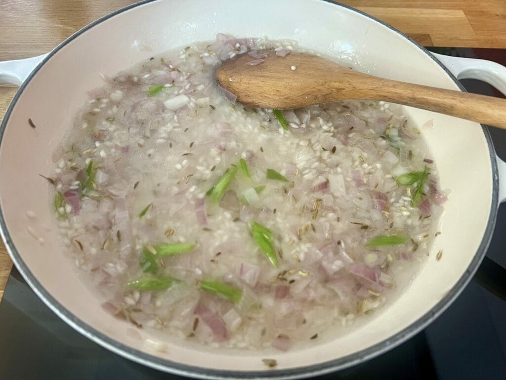 A wooden spoon stirs chopped onions, green chilies, cumin seeds, and garlic simmering in oil inside a white pot on a stovetop.