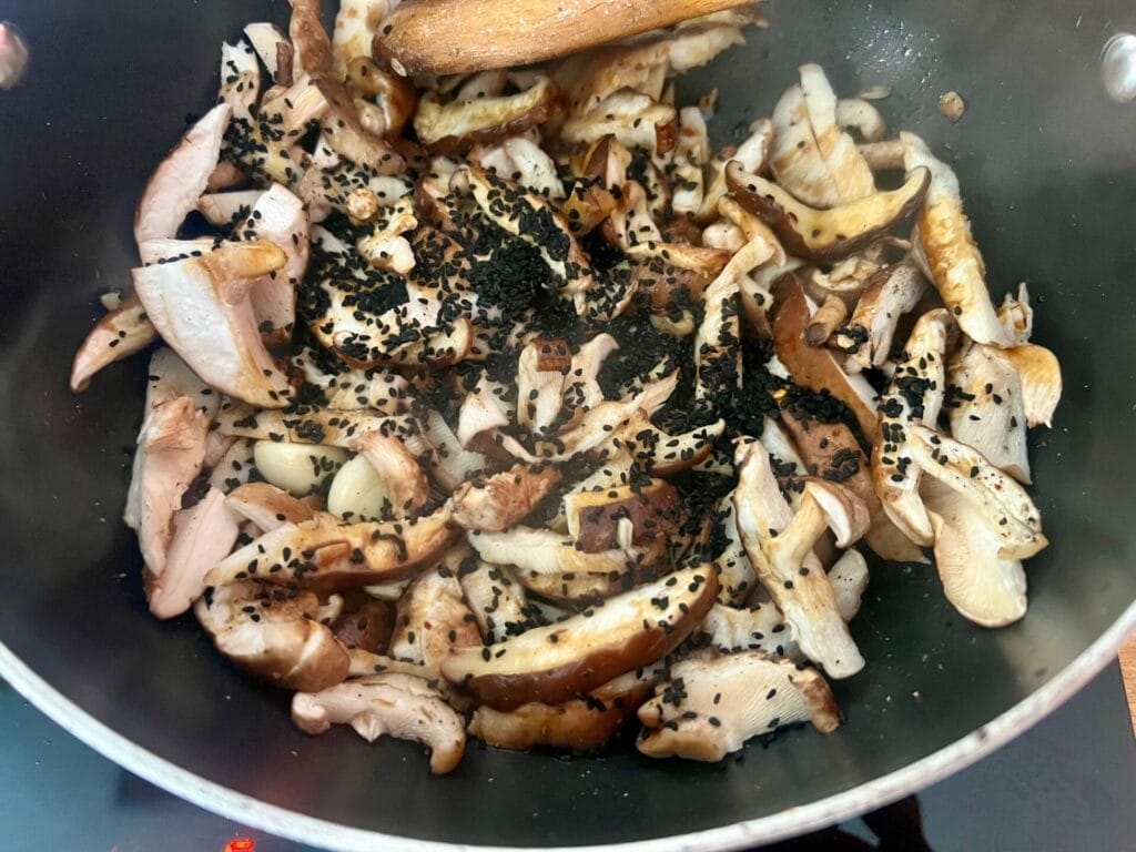 A pan filled with chopped mushrooms and black cumin seeds being cooked, stirred with a wooden spoon.