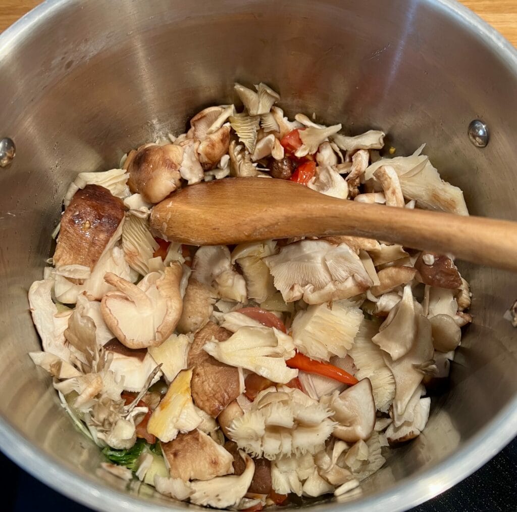 A wooden spoon stirs a variety of sliced mushrooms, red peppers, and other vegetables in a silver pot, creating a fragrant Anti-inflammatory Shiitake Mushroom and Lemongrass Soup for Immune Support.
