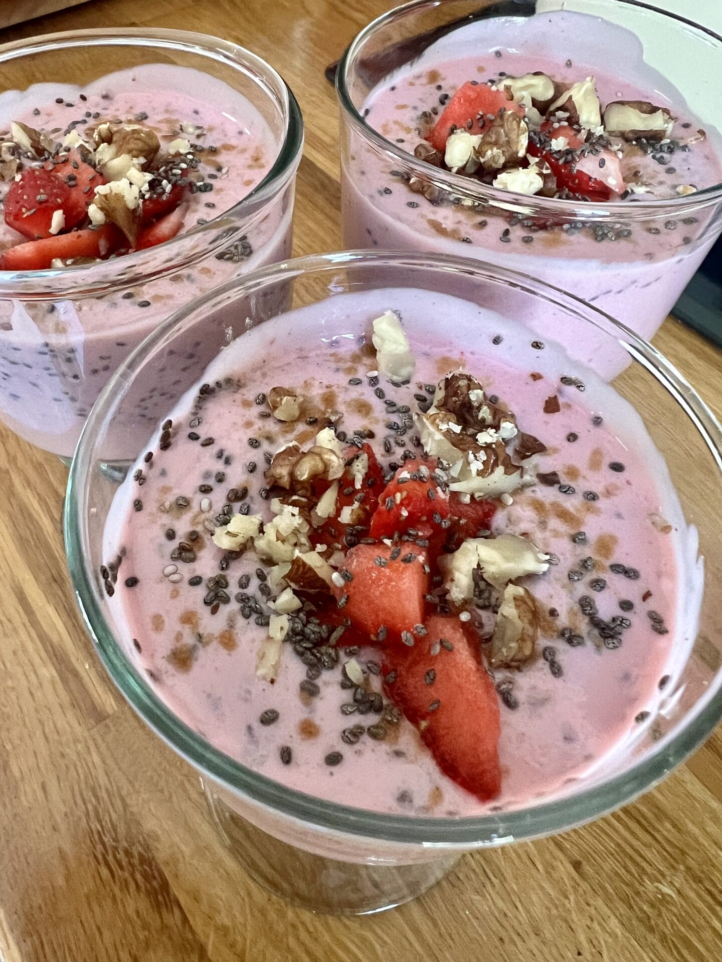 Three glass containers filled with pink smoothie topped with chia seeds, chopped nuts, and pieces of strawberries on a wooden surface. Three glass containers filled with pink smoothie topped with chia seeds, chopped nuts, and pieces of strawberries on a wooden surface.