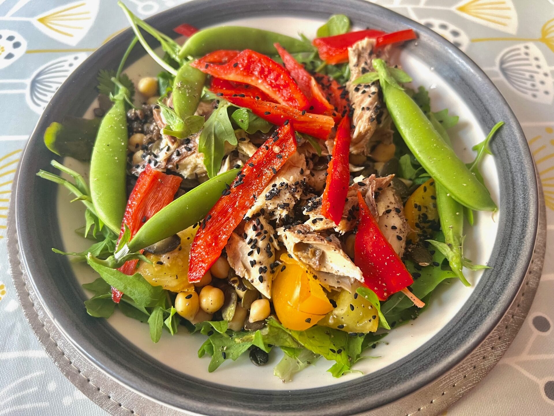 A bowl of green salad topped with slices of red bell pepper, yellow tomatoes, snap peas, chickpeas, sunflower seeds, and what appears to be shredded chicken, placed on a decorative tablecloth.