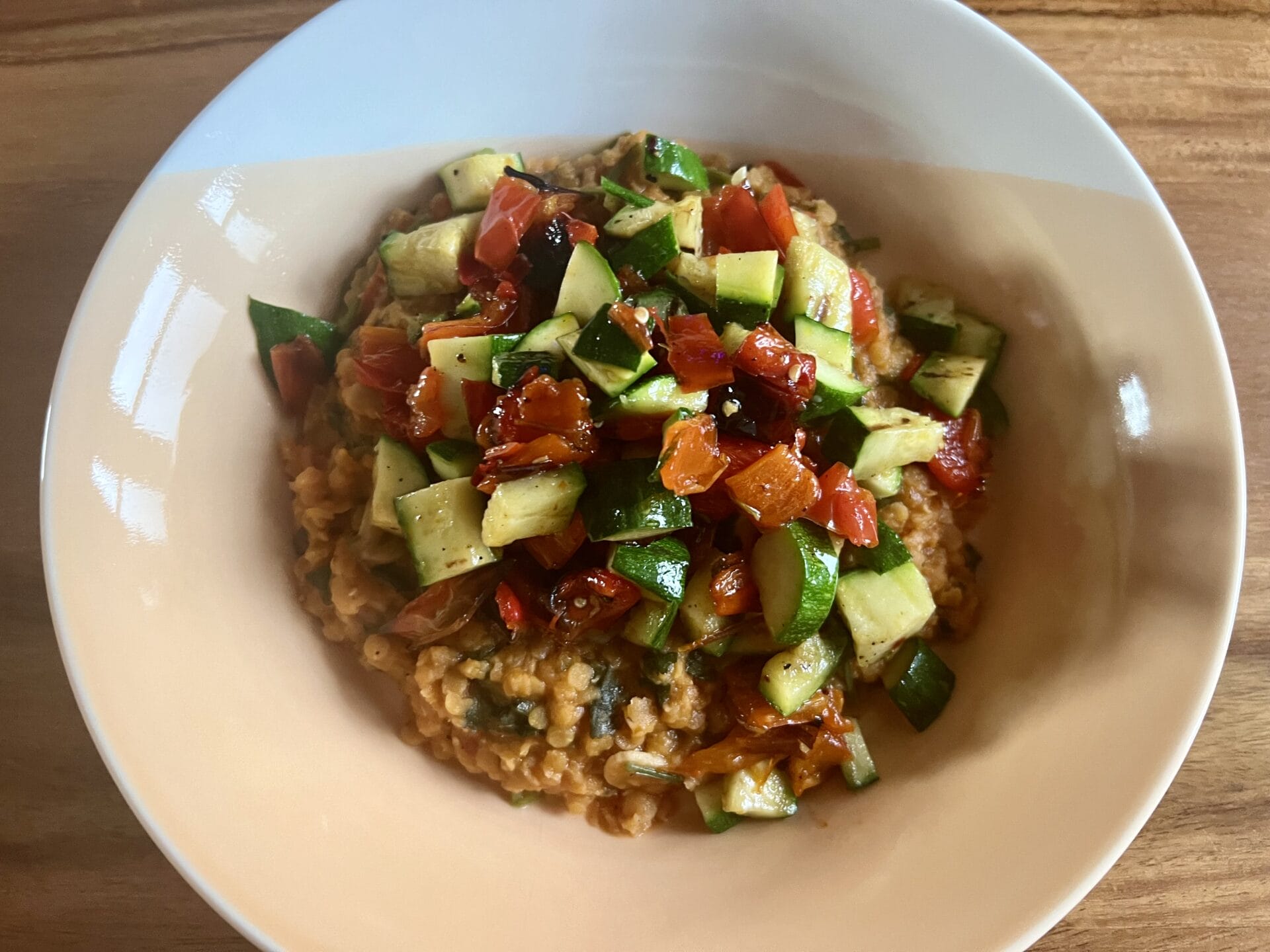 A bowl of dhal topped with chopped vegetables including tomatoes, courgettes and green bell peppers, served on a wooden table.