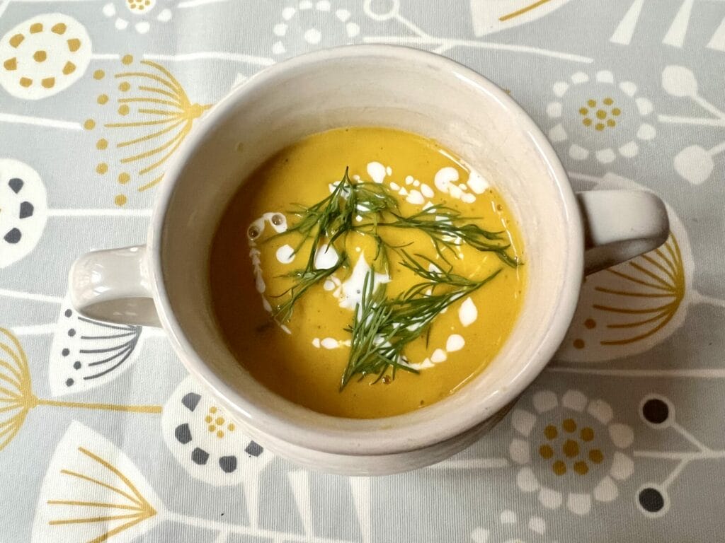 A bowl of creamy Anti-Inflammatory Butternut Squash and Carrot Soup with swirls of cream and green herbs, including a hint of turmeric ginger and dill, placed on a floral-patterned tablecloth.