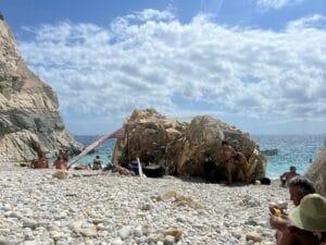 Rocky beach with several people sunbathing and relaxing. A makeshift shelter is built against large rocks, and the sea is visible in the background under a partly cloudy sky.