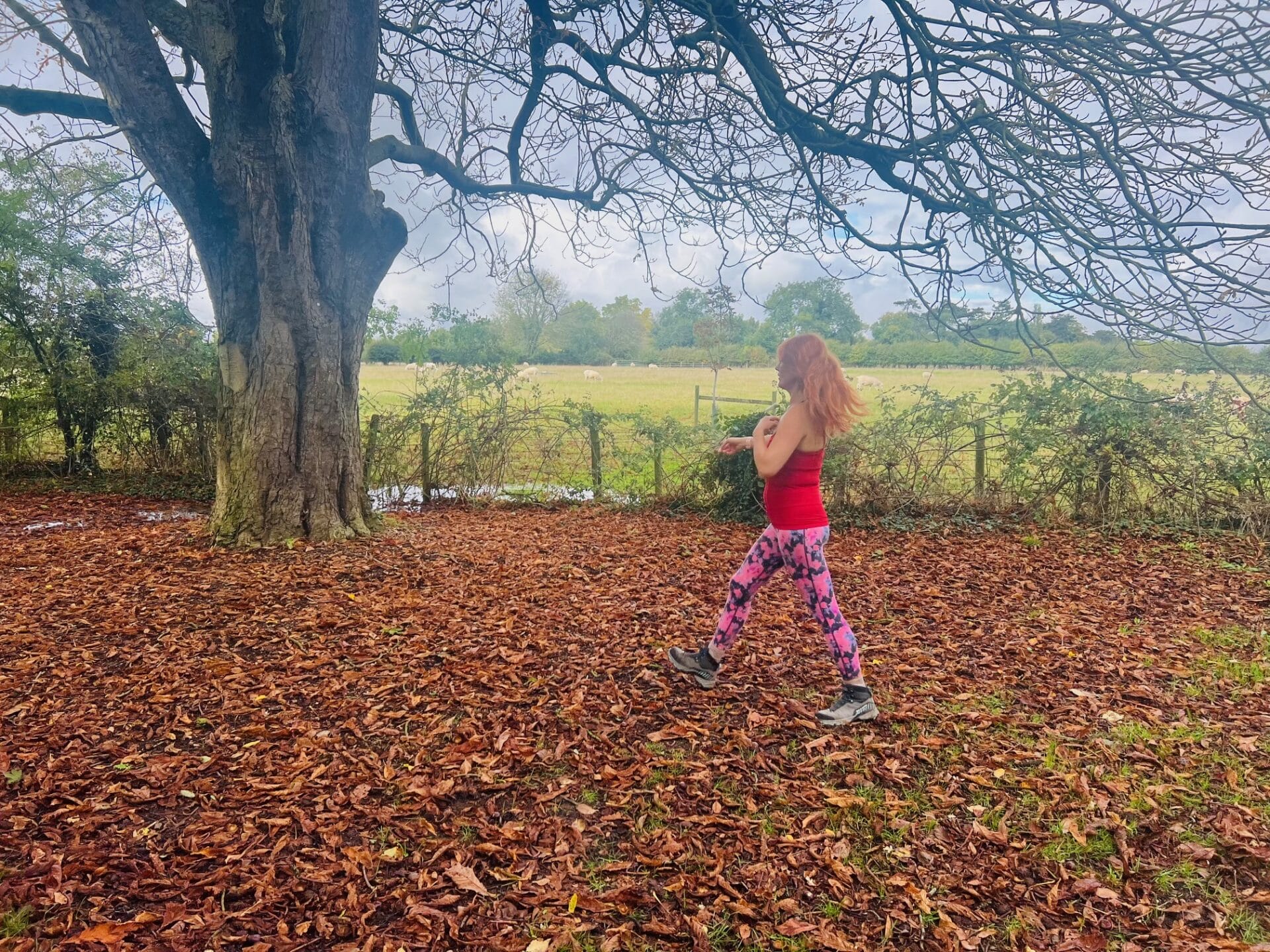 A person with long red hair strolls along a leaf-covered path, wearing a red top and patterned leggings, embracing the autumnal tune for wellbeing beside a large tree in the serene countryside.