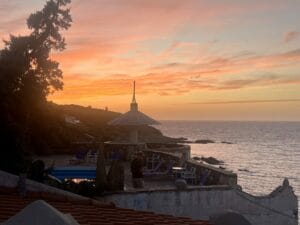 Sunset view over an ocean with a patio in the foreground, featuring lounge chairs and a partial roof structure. A person stands near the railing, experiencing tranquility at the Ikaria Yoga Retreat, as distant land is silhouetted against the sky.