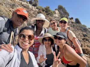 A group of seven people, soaking up the sun at the Ikaria Yoga Retreat, casually dressed in outdoor attire and hats, pose for a selfie amidst the rocky, hilly landscape. Two of them flash peace signs, capturing the spirit of their rejuvenating escape.