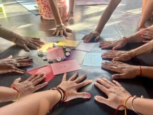 At the Ikaria Yoga Retreat, a group of people with colorful nail polish and bracelets places their hands on a table surrounding cards and stones arranged in a circle, creating a serene atmosphere of mindfulness and connection.