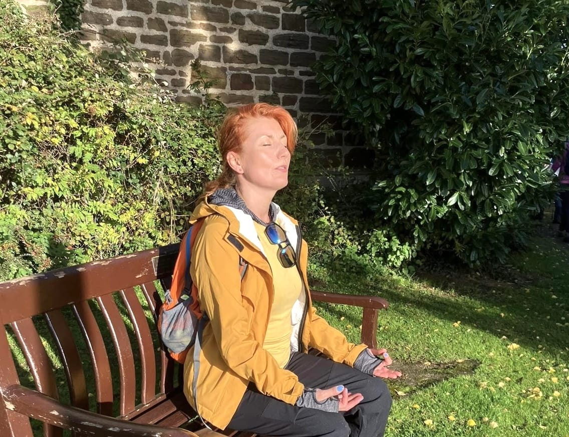 A person with red hair sits on a wooden bench in a garden, wearing a yellow jacket and backpack, meditating with eyes closed, breathing deeply and consciously supporting their lungs and immune system.