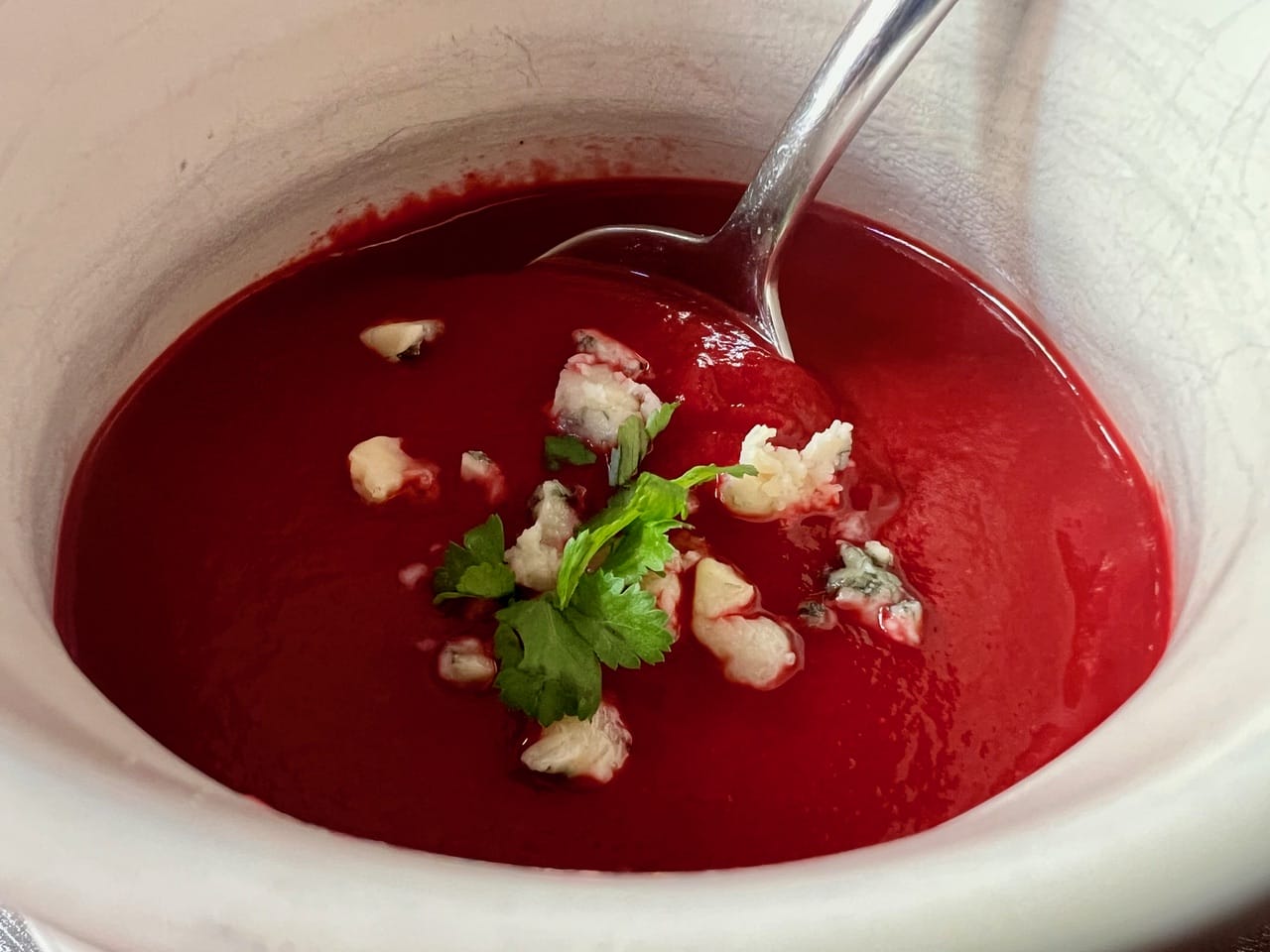 A bowl of red soup with a spoon. Topped with cream, blue cheese crumbles, and a sprig of cilantro.