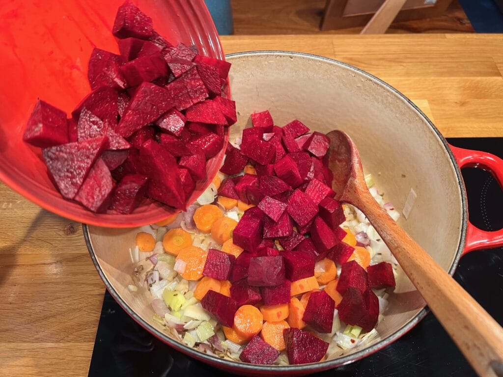 Red beets are being added to a pot containing chopped onions and carrots with a wooden spoon on the side.
