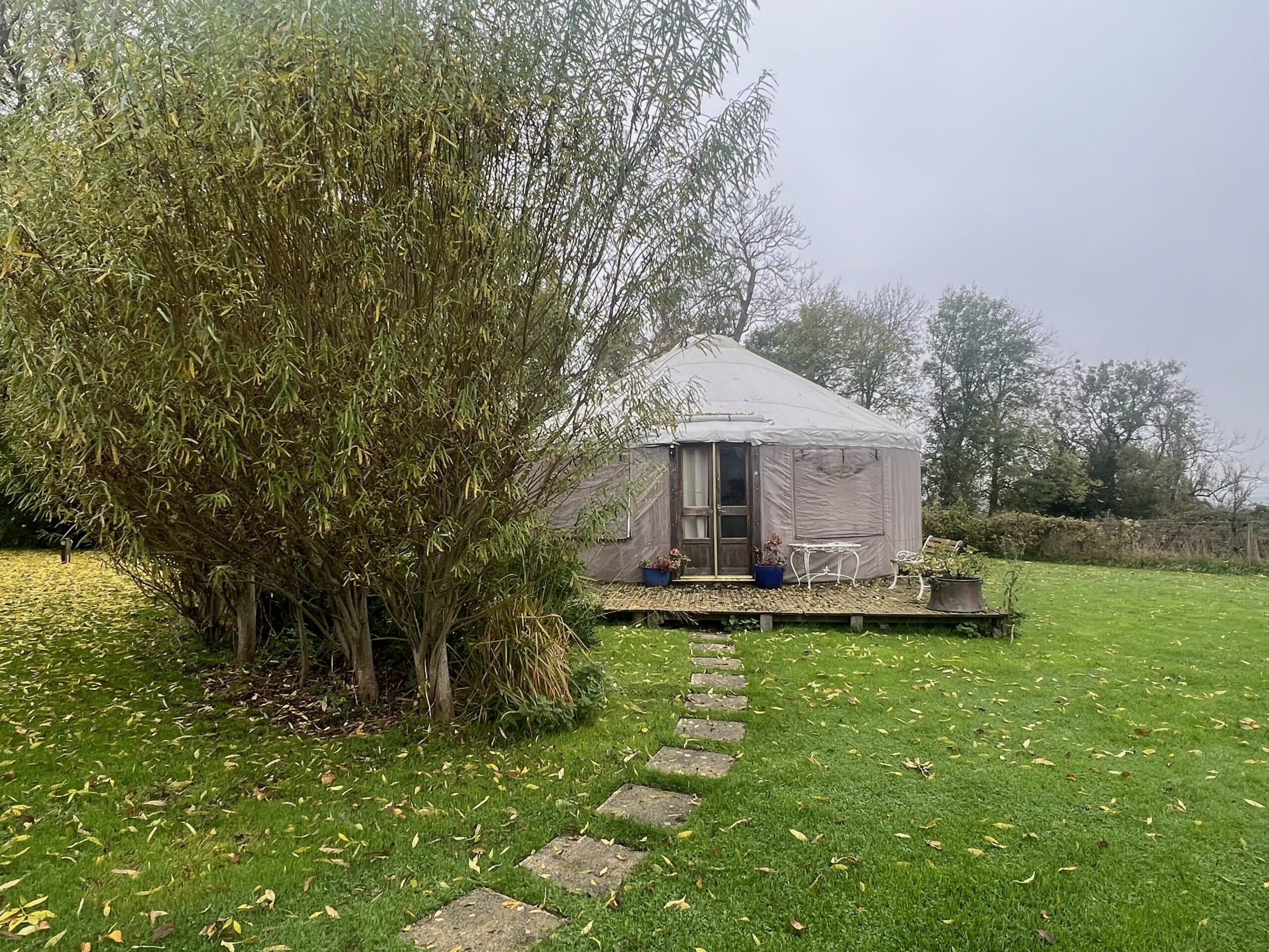 A round yurt with a white roof sits on a wooden deck, surrounded by green grass and trees. A stone path leads to the entrance. A round yurt with a white roof sits on a wooden deck, surrounded by green grass and trees. A stone path leads to the entrance.