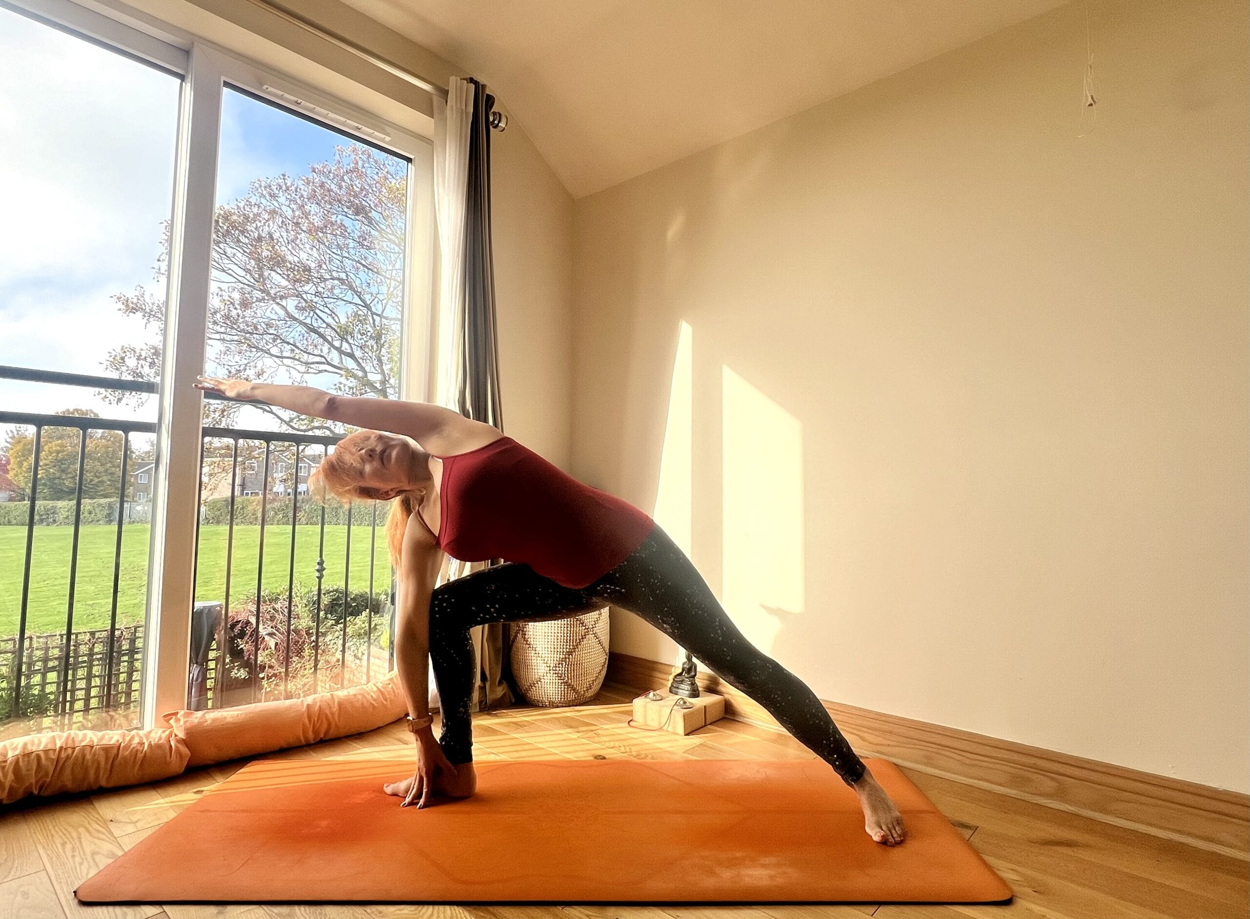 A person practices a gentle yoga pose on an orange mat in a sunlit room with a large window overlooking a grassy area, blending warming poses to keep the body balanced—even on cooler days. A person practices a gentle yoga pose on an orange mat in a sunlit room with a large window overlooking a grassy area, blending warming poses to keep the body balanced—even on cooler days.