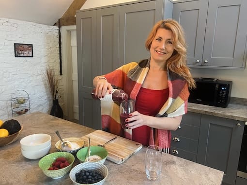 Woman pouring juice into a glass in a kitchen. Various fruits and bowls are on the counter.