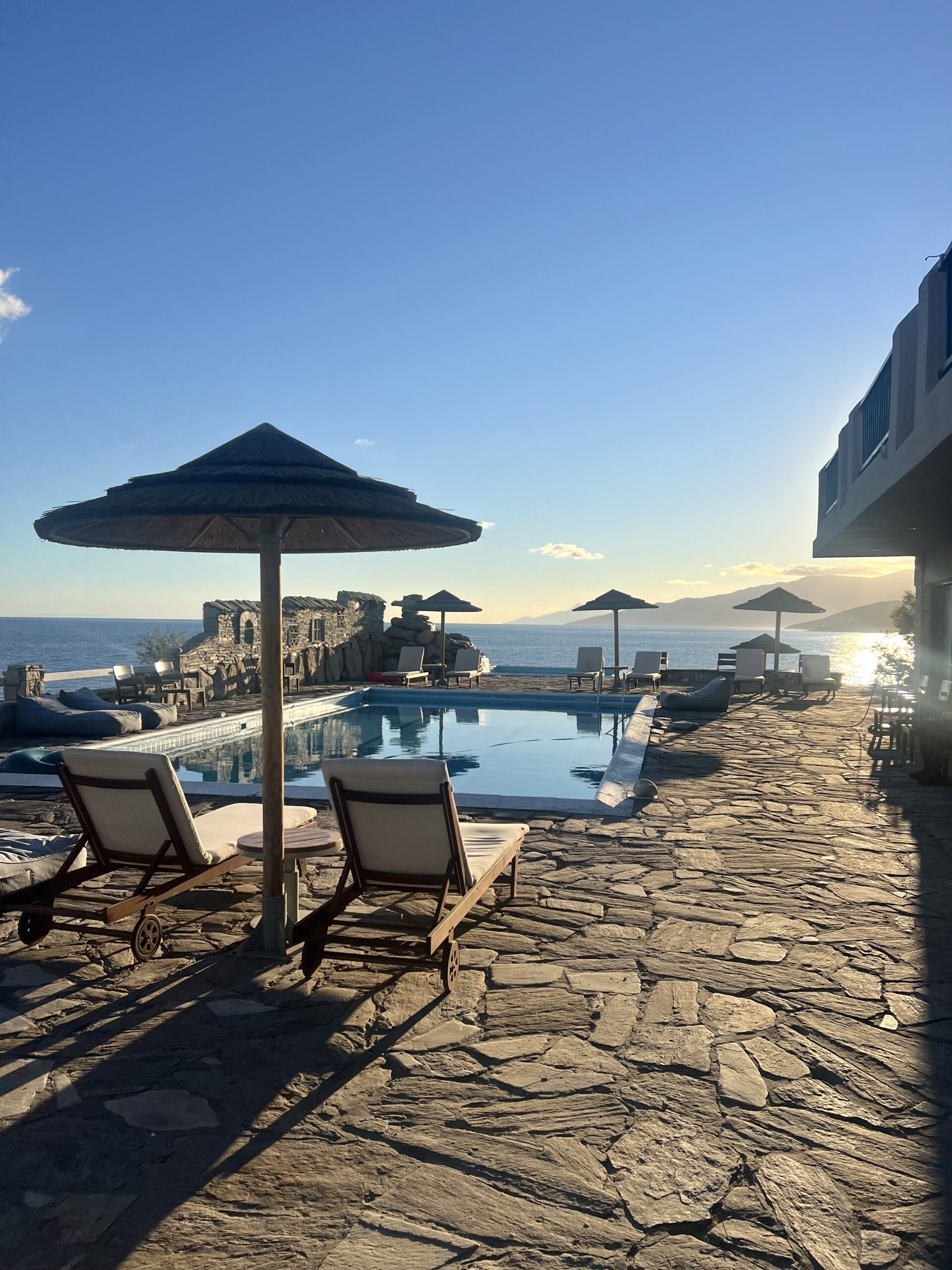 Sunlit poolside scene with empty lounge chairs and umbrellas, overlooking a calm sea and distant mountains under a clear blue sky.