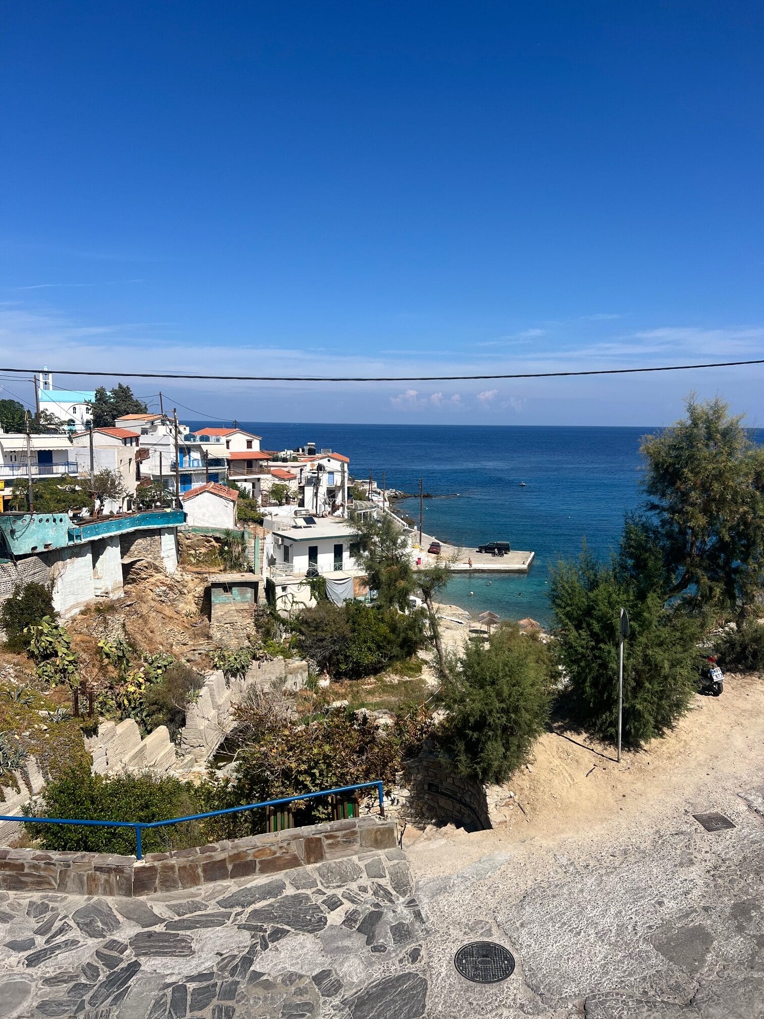 Seaside village with white buildings and lush greenery on a hill, overlooking the blue ocean under a clear sky.