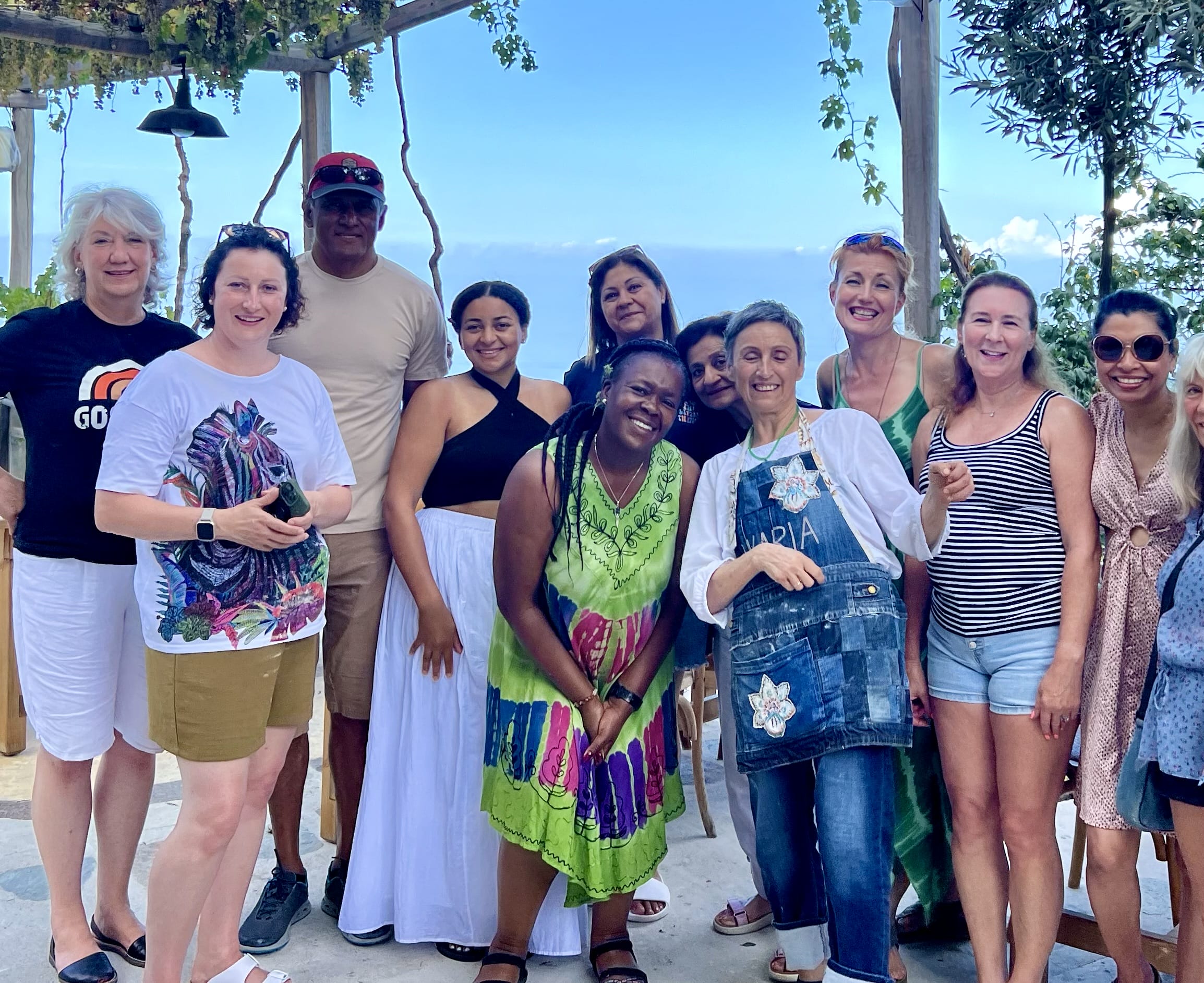 Group of people smiling and posing outdoors under a pergola with a scenic background of trees and sea.