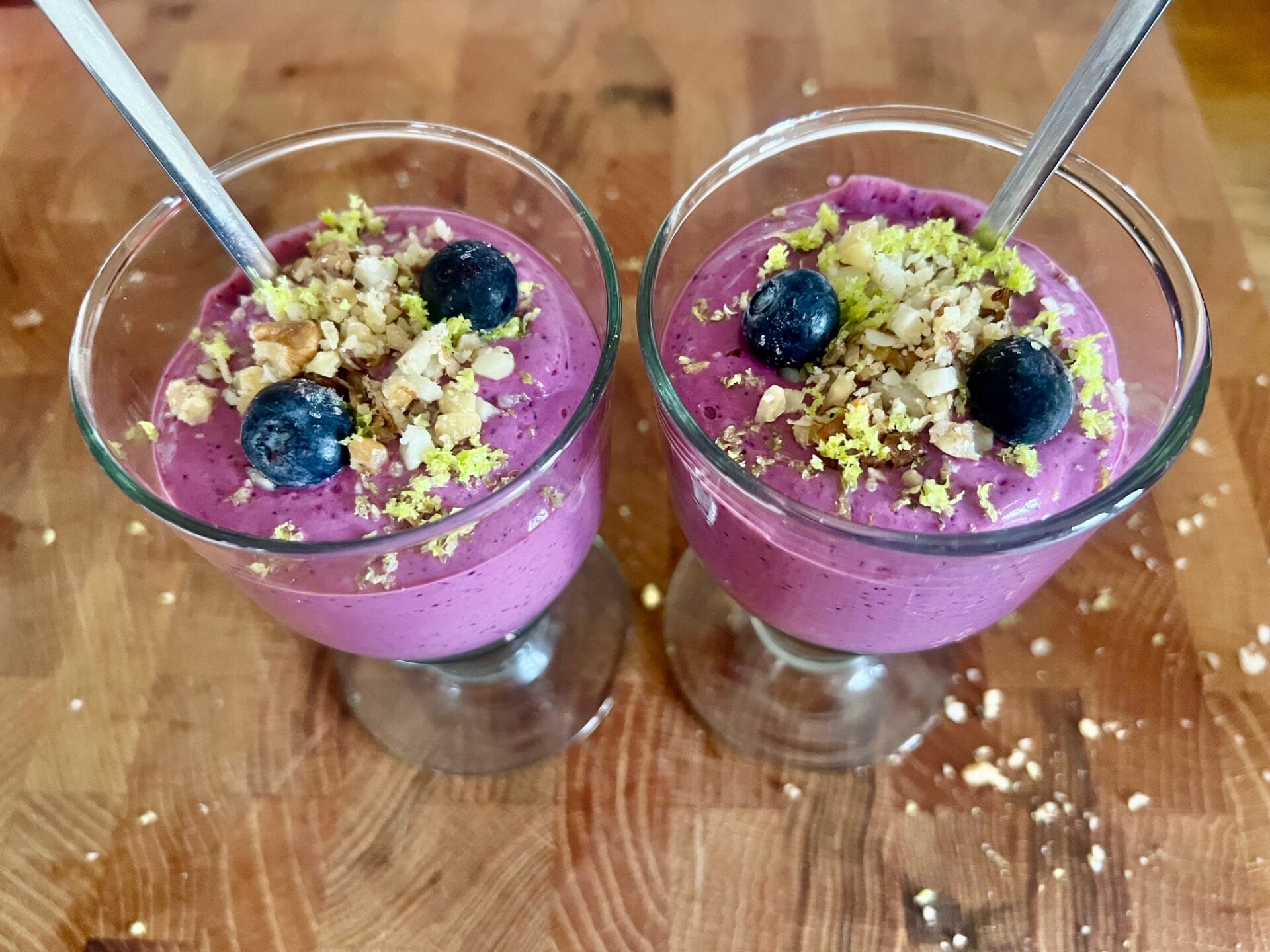 Two glass cups filled with Velvet Berry Mousse, topped with blueberries, crushed nuts, and grated zest, each with a spoon, on a wooden surface.
