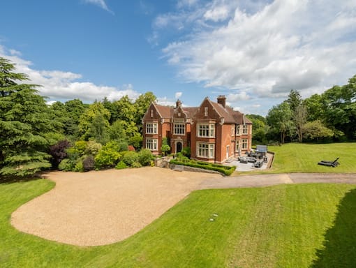A large red brick house with a gravel driveway, surrounded by lush greenery and trees under a blue sky.
