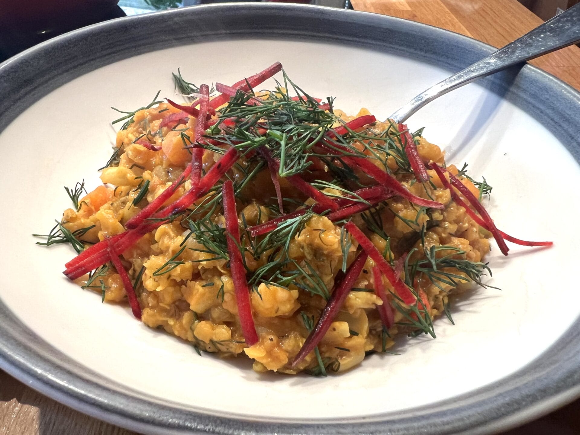 A bowl of dhal with fresh dill and delicate red beet sticks, presented in a blue-rimmed dish alongside a spoon, reminiscent of a vibrant red lentil dhal with roasted beetroots.