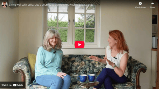 Two women sit on a sofa talking in a room with a window. There are two mugs and a small table with water bottles between them.