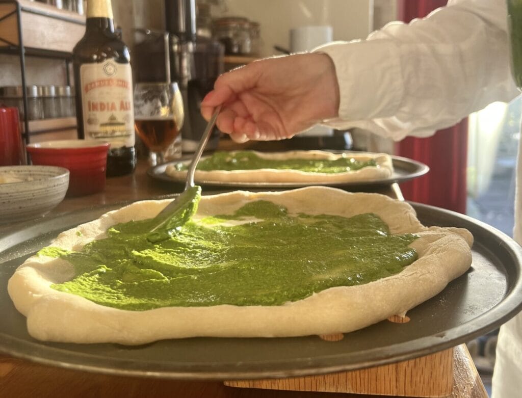 Person spreading green sauce on pizza dough in a kitchen, with a bottle of India Pale Ale and a glass nearby.