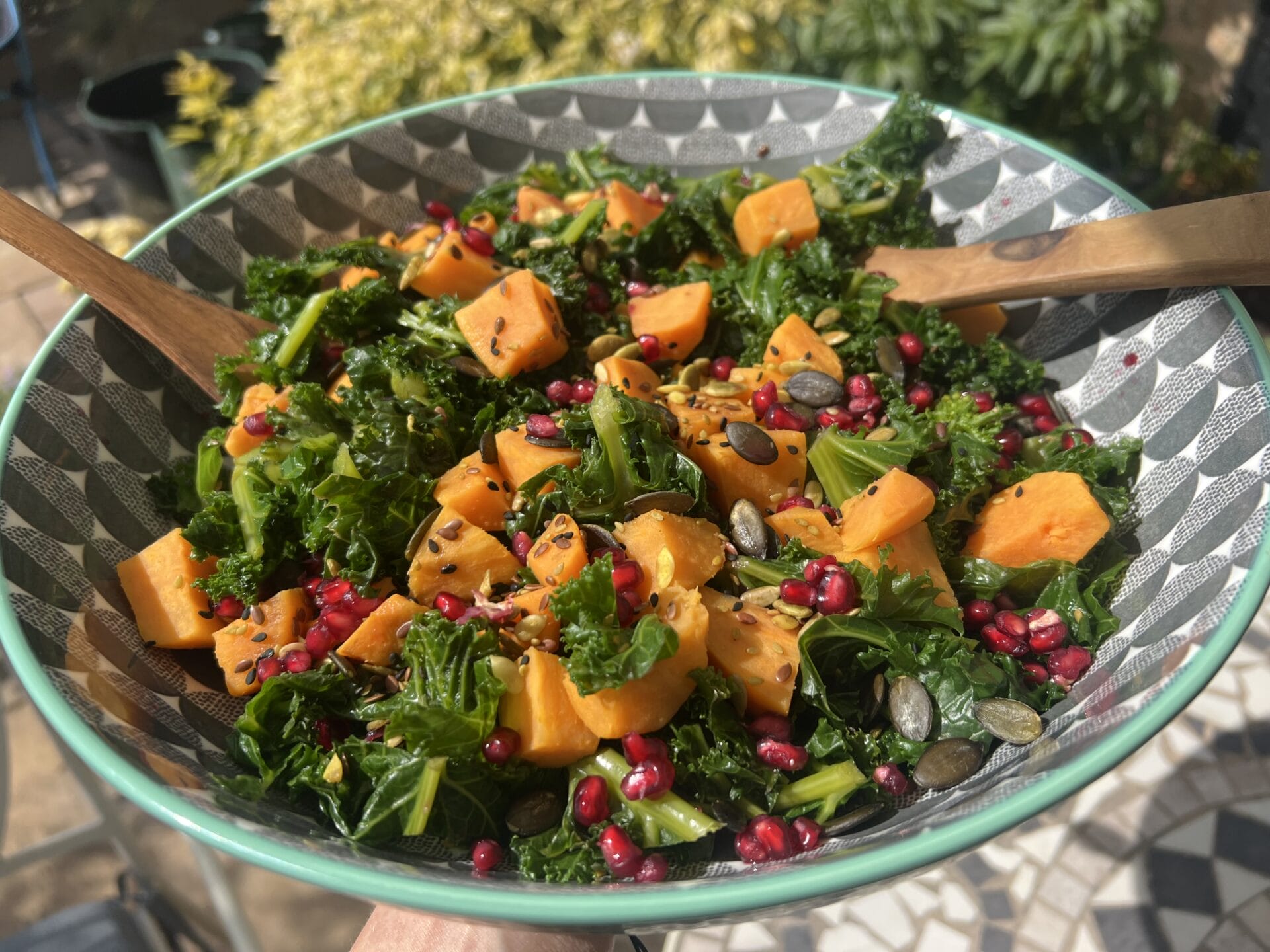 A salad with kale, sweet potato chunks, pomegranate seeds, and pumpkin seeds in a patterned bowl, mixed with wooden utensils. A salad with kale, sweet potato chunks, pomegranate seeds, and pumpkin seeds in a patterned bowl, mixed with wooden utensils.
