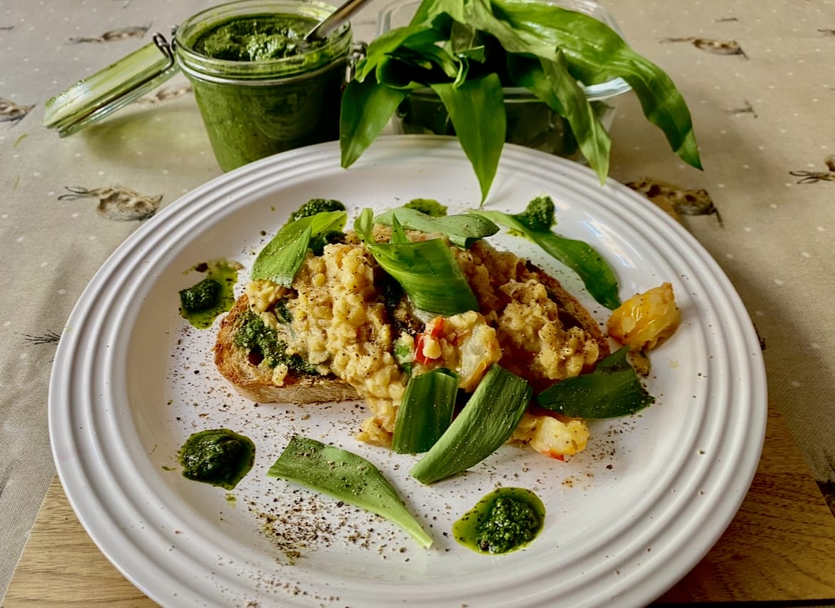 A plate of scrambled eggs on toast with green leaves and wild garlic pesto, garnished with herbs and pepper. A jar of pesto and greens are in the background.