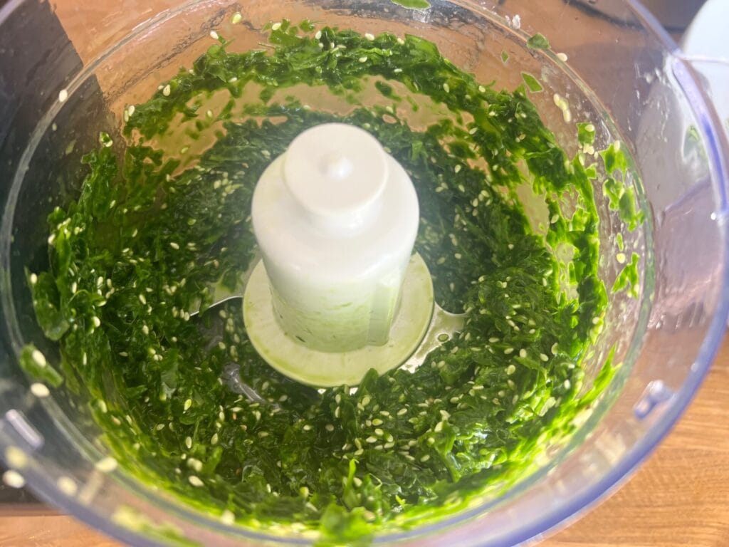 Close-up of a food processor containing freshly chopped green herbs for Green Detox Delight: Wild Garlic Pancakes with Mediterranean Garnish, with visible seeds and small pieces.
