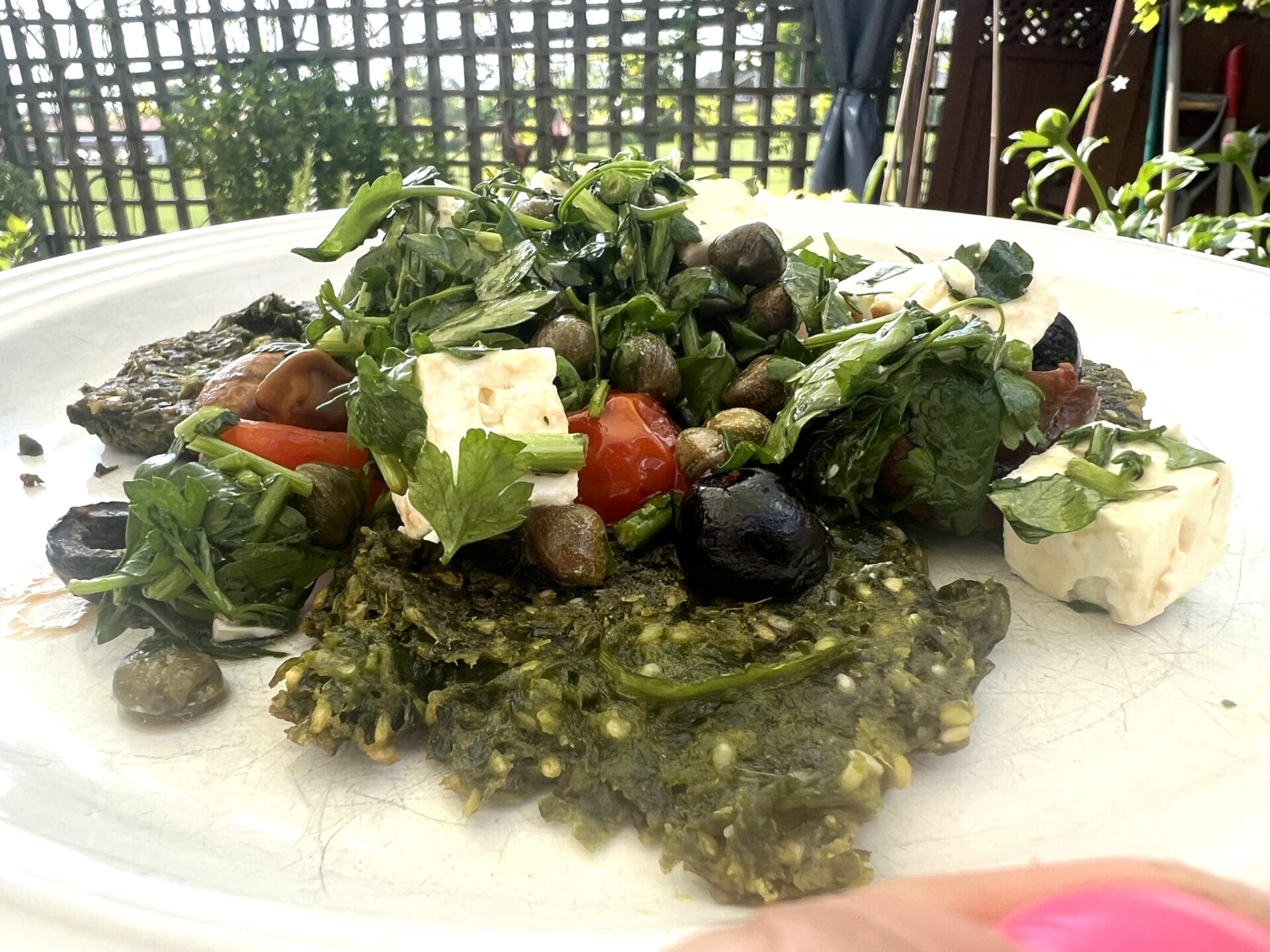 A plate of Green Detox Delight: wild garlic pancakes with Mediterranean garnish—patties topped with arugula, cherry tomatoes, olives, capers, and feta cubes—set outdoors on a sunny day.