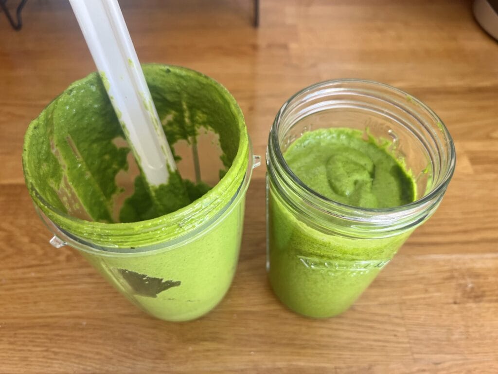 Two containers of a vibrant green smoothie grace the wooden surface; one is a blender cup with blades, and the other is a filled glass jar, reminiscent of fresh Wild Garlic Pesto.