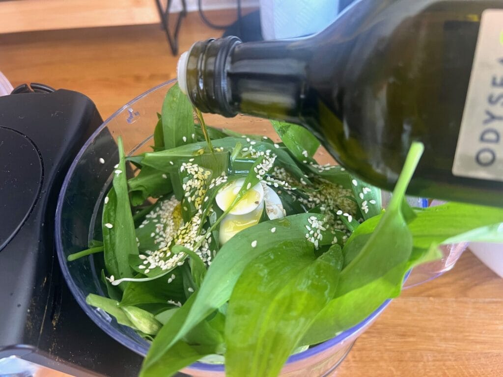 Olive oil is being poured into a food processor containing fresh green leaves and sesame seeds to craft a Green Detox Delight: Wild Garlic Pancakes with Mediterranean Garnish sauce or dressing.