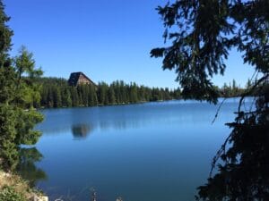 A calm lake surrounded by evergreen trees, with a large triangular-roofed building in the distance under a clear blue sky.