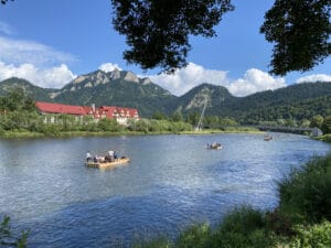 Several wooden rafts with people float on a wide river, surrounded by green hills and mountains under a blue sky with scattered clouds. A building with a red roof is visible on the riverbank.
