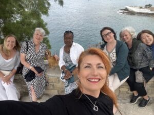Six women sit together on a stone wall by the water, smiling at the camera; a small brown dog is lying near them. Trees and the sea are visible in the background.