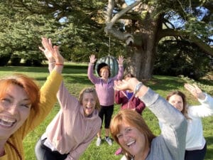 Five women pose outdoors, smiling and waving at the camera in front of a large tree and a tire swing on a sunny day.