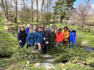 A group of thirteen women poses outdoors in front of a pond and trees on a sunny day, wearing coats and jackets.