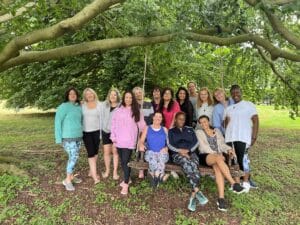 A group of fourteen women pose together outdoors under a large tree, with two sitting on a wooden swing and the others standing behind them.