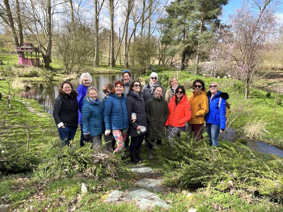 A group of thirteen women poses outdoors in front of a pond and trees on a sunny day, wearing coats and jackets.